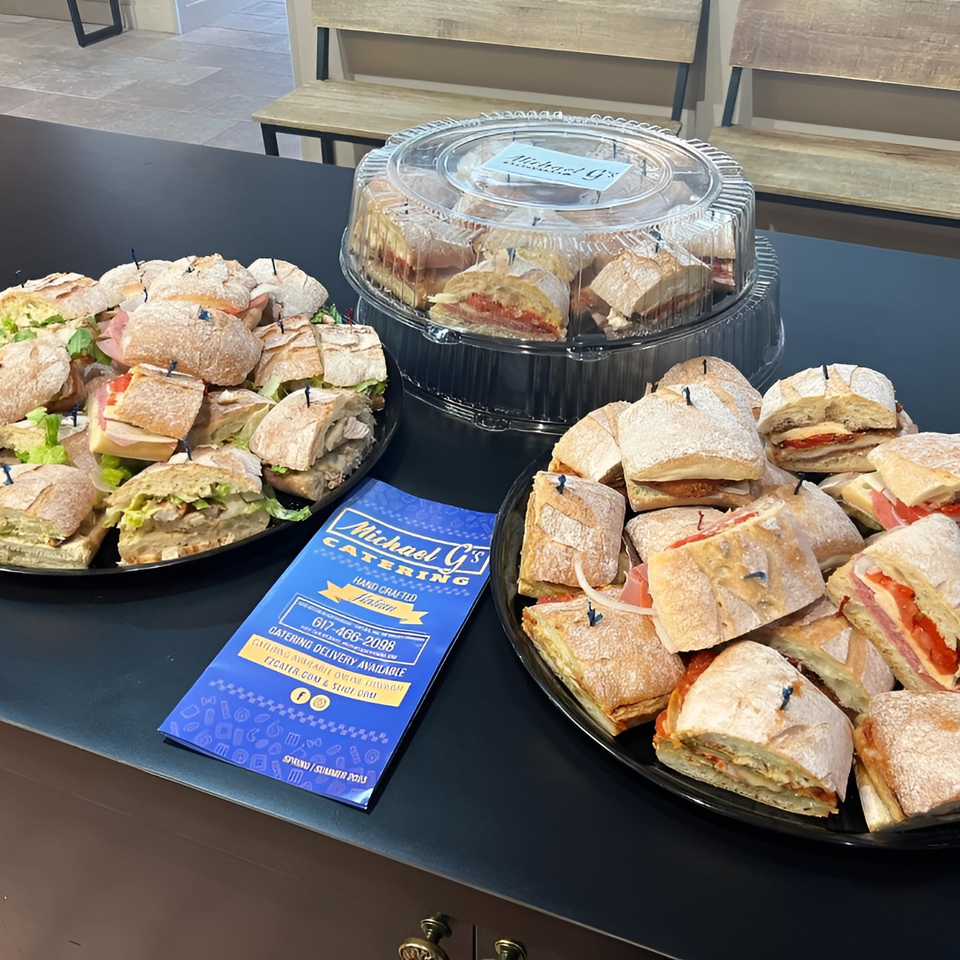 Michael G's catering display with platters of Italian sandwiches and a brochure on a black table.